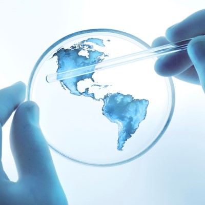 A map of the world is shown in blue liquid on a petri dish. The dish is held by a gloved hand. A second gloved hand holds a pipette.